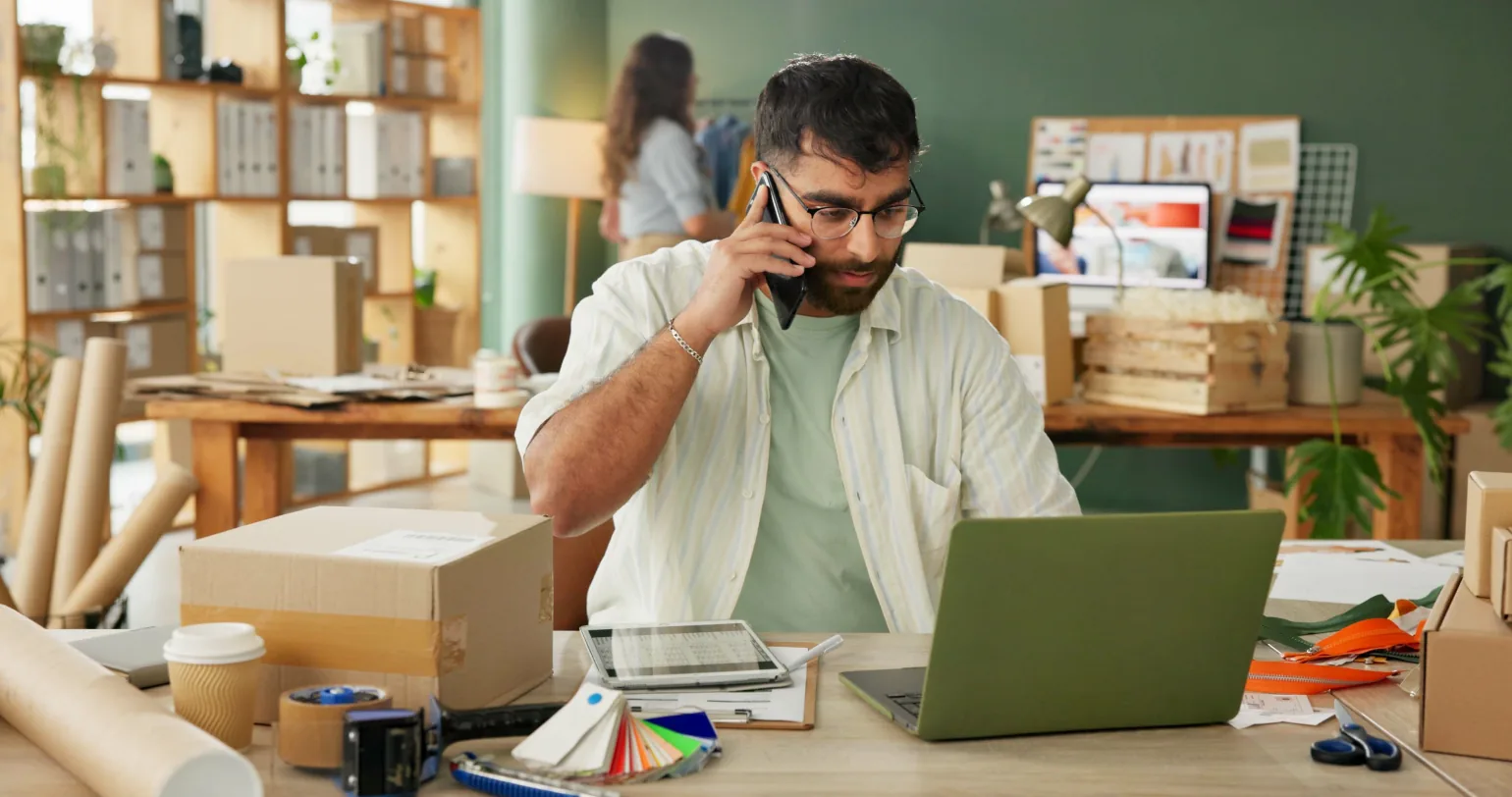 A print-on-demand store owner taking a call at his packing desk, laptop open beside shipping boxes and colour swatches